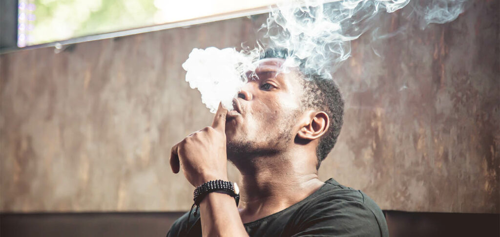 Portrait of a black young man smoking and playing with the smoke from a water pipe while is sitting in a sofa inside a room with a window